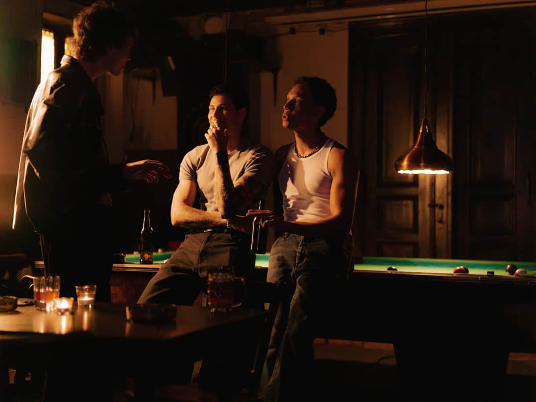 Three people talking at a pool table in a dimly lit dive bar with pendant lights