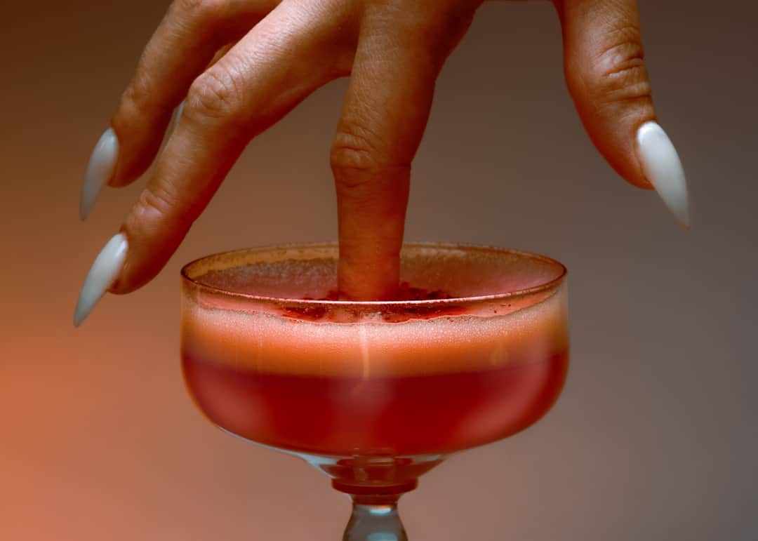 Hand dipping into red cocktail in coupe glass