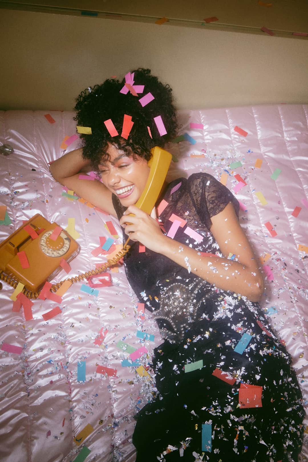 Woman lying on confetti-covered bed with colorful 80s-style hair clips and sequined black top in vibrant celebration scene