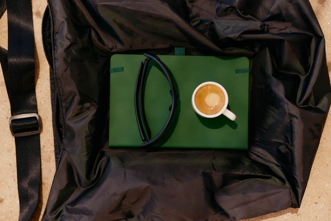 Black eyewear frames on a green laptop case with a latte coffee cup on black fabric