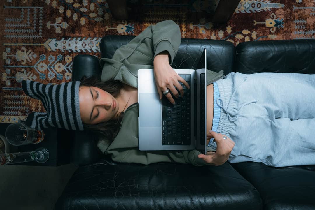 Woman working from home on laptop while lying on black leather sofa