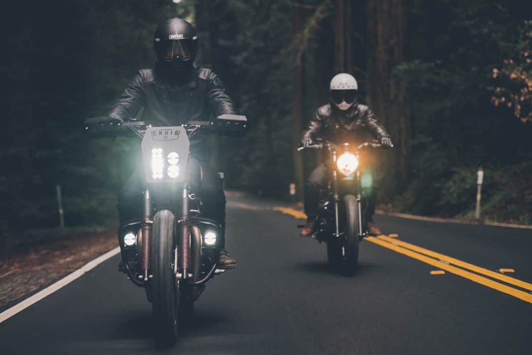Two motorcyclists speeding through a dark forest road with bright headlights at night