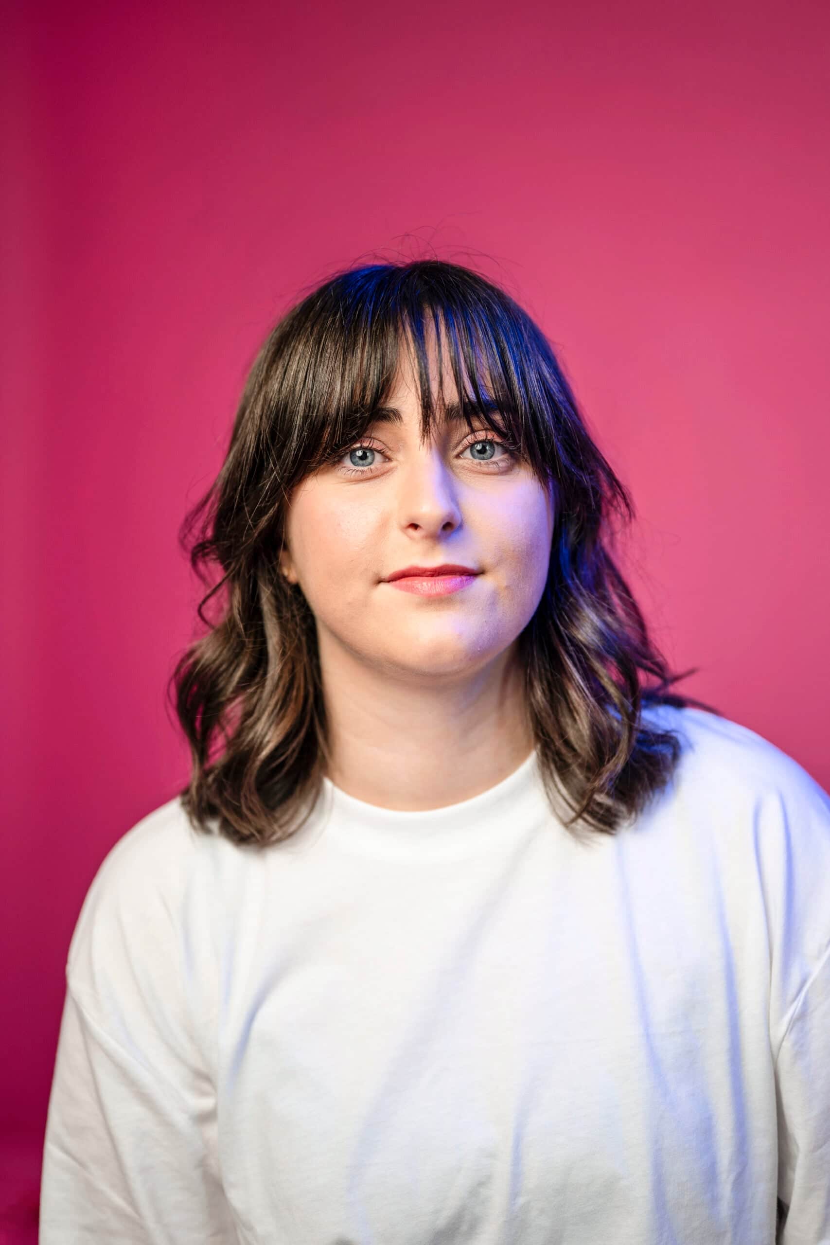 Woman with dark wavy hair and bangs wearing white shirt against pink background, professional headshot