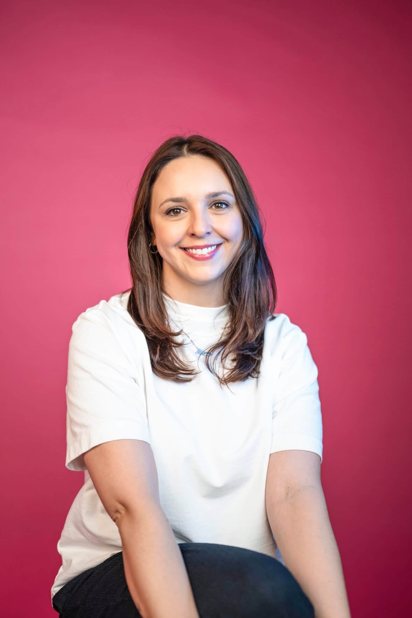 Professional marketing team member with dark hair in studio headshot wearing white shirt against pink background