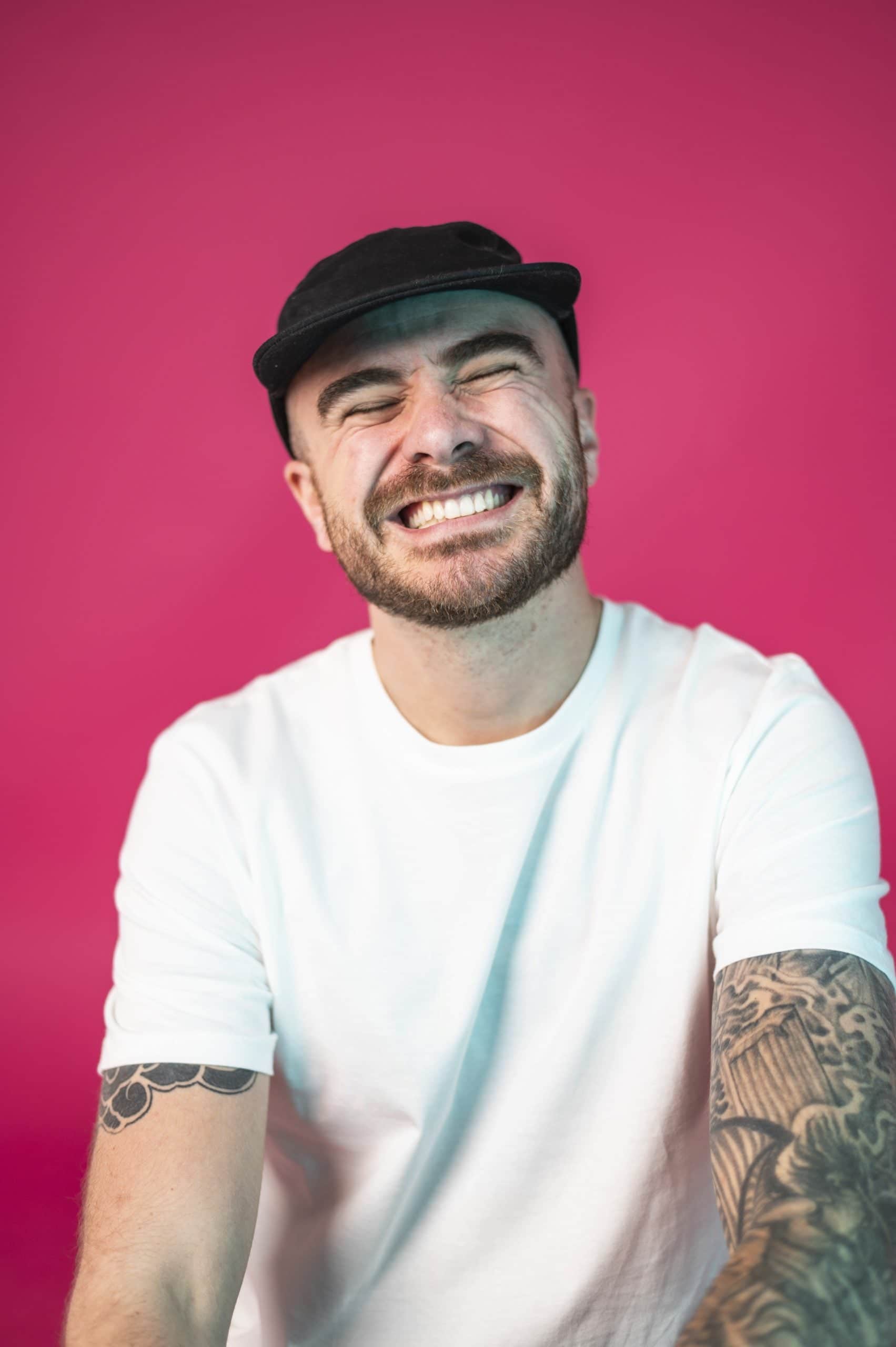 Man smiling broadly, wearing a white t-shirt and black cap, tattooed arm, against a vibrant pink background.