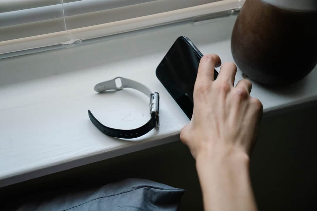 From above of crop anonymous person taking cellphone placed on windowsill near smart watch while waking up in morning (Photo: Eren Li via Pexels)