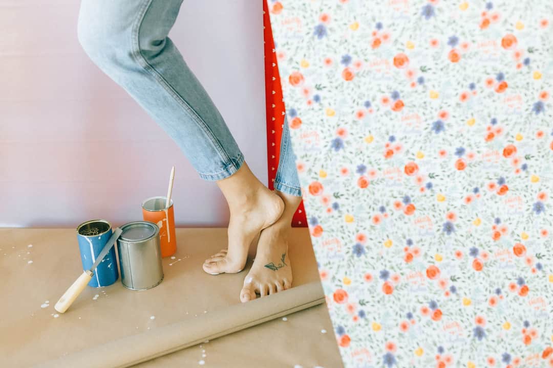 A woman decorates a room with floral wallpaper, surrounded by paint cans and a brush. (Photo: Nataliya Vaitkevich via Pexels)