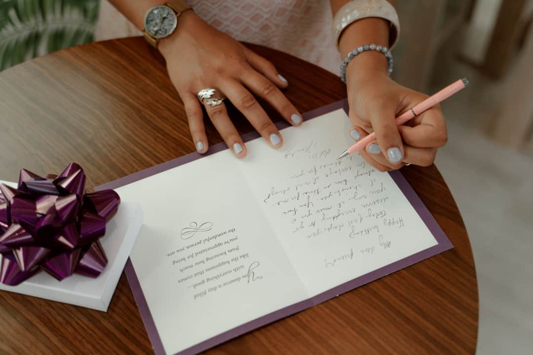 A woman writes a message inside a greeting card on a wooden table with a gift beside. (Photo: RDNE Stock project via Pexels)