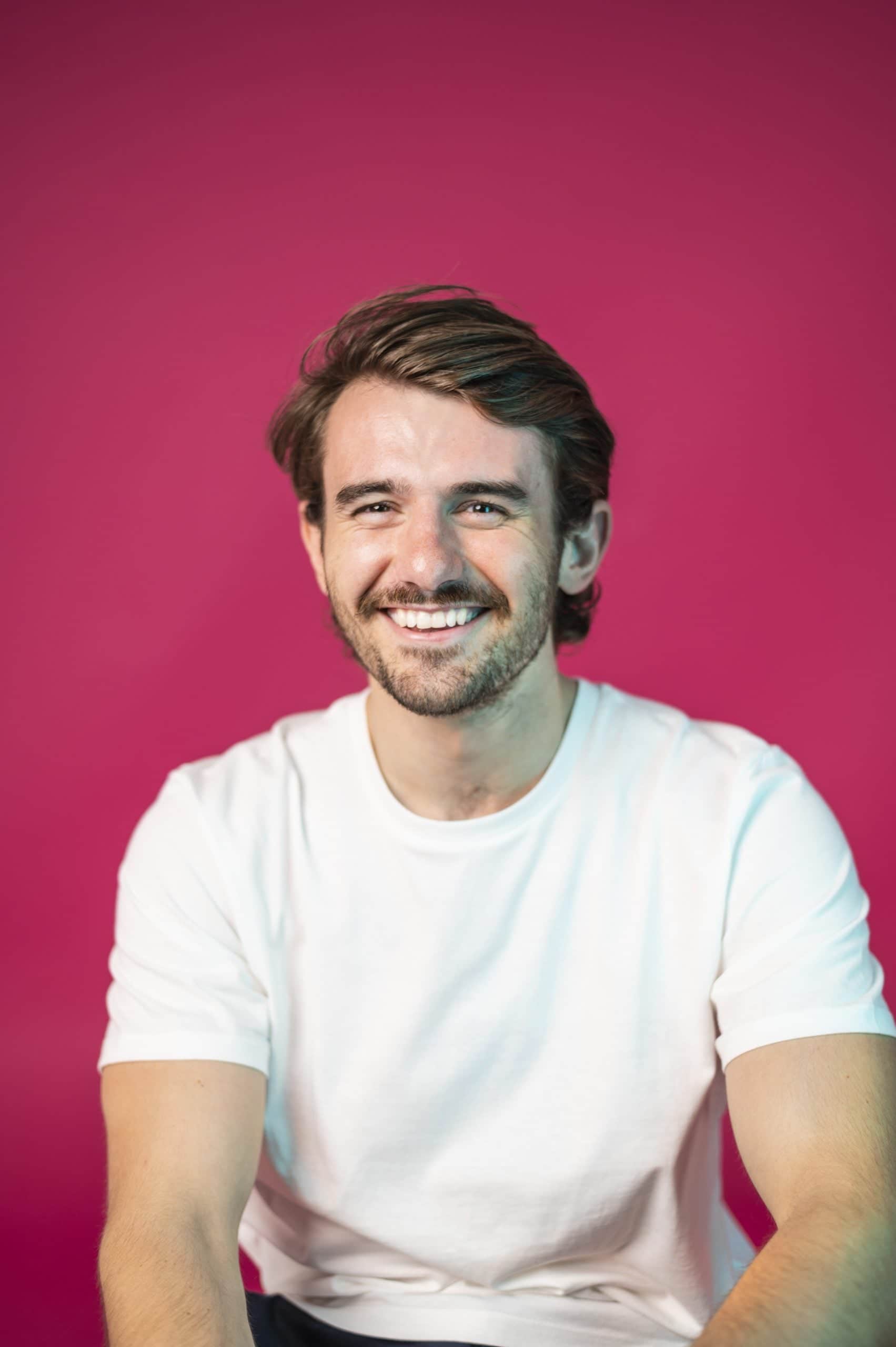 Person smiling, wearing a white t-shirt against a vibrant pink background, representing friendly brand image.