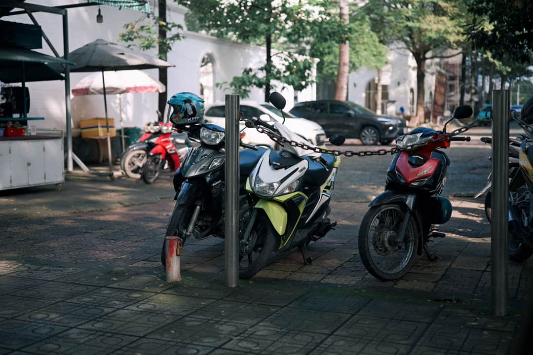 Three motorcycles chained together on a city sidewalk, shaded by trees and near a food stall. (Photo: 🇻🇳🇻🇳 Việt Anh Nguyễn 🇻🇳🇻🇳 via Pexels)