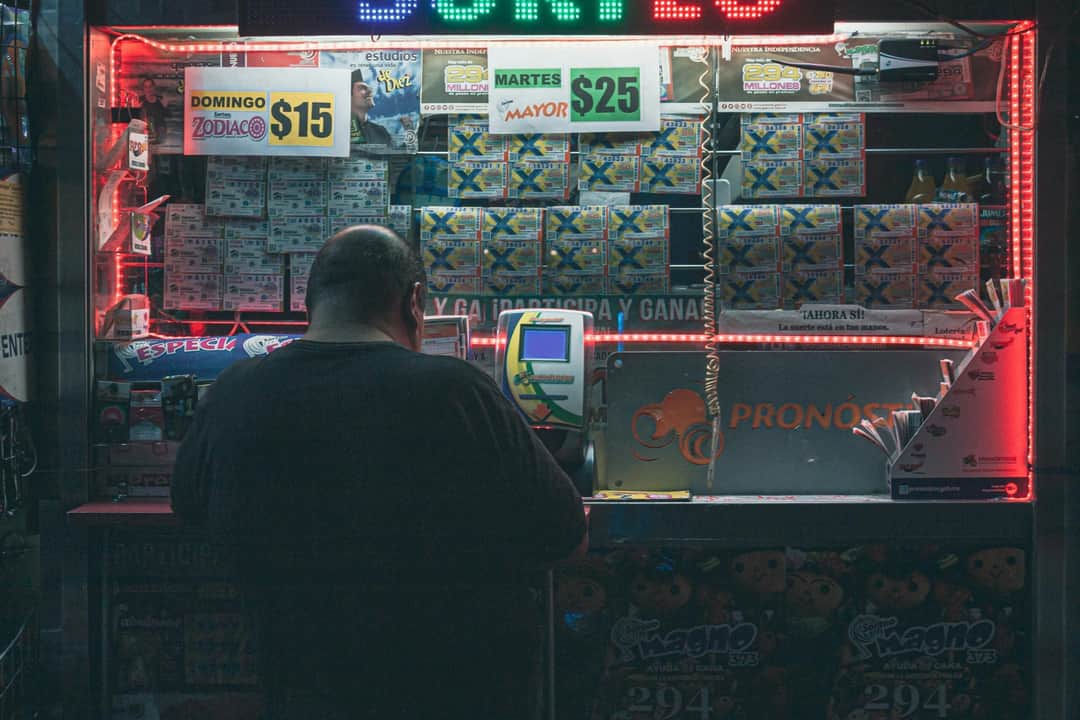 A man purchasing a lottery ticket at an illuminated kiosk in Mexico City, highlighting risk and hope. (Photo: Peaton Hugo via Pexels)
