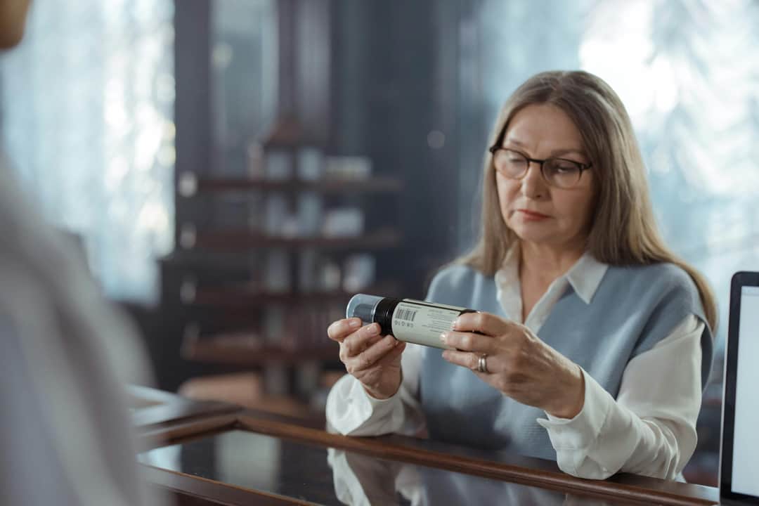 A senior woman with eyeglasses reviews a medicine bottle at a pharmacy counter indoors. (Photo: cottonbro studio via Pexels)
