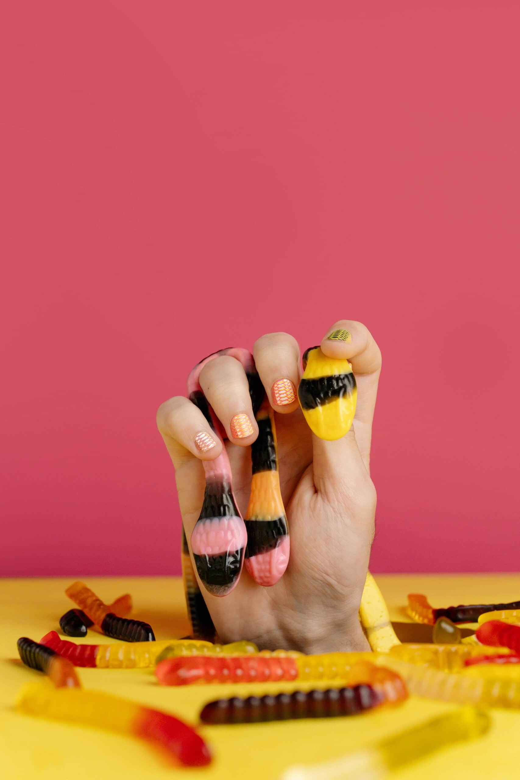 Hand holding gummy worm candies in creative candy arrangement with vibrant pink and yellow background