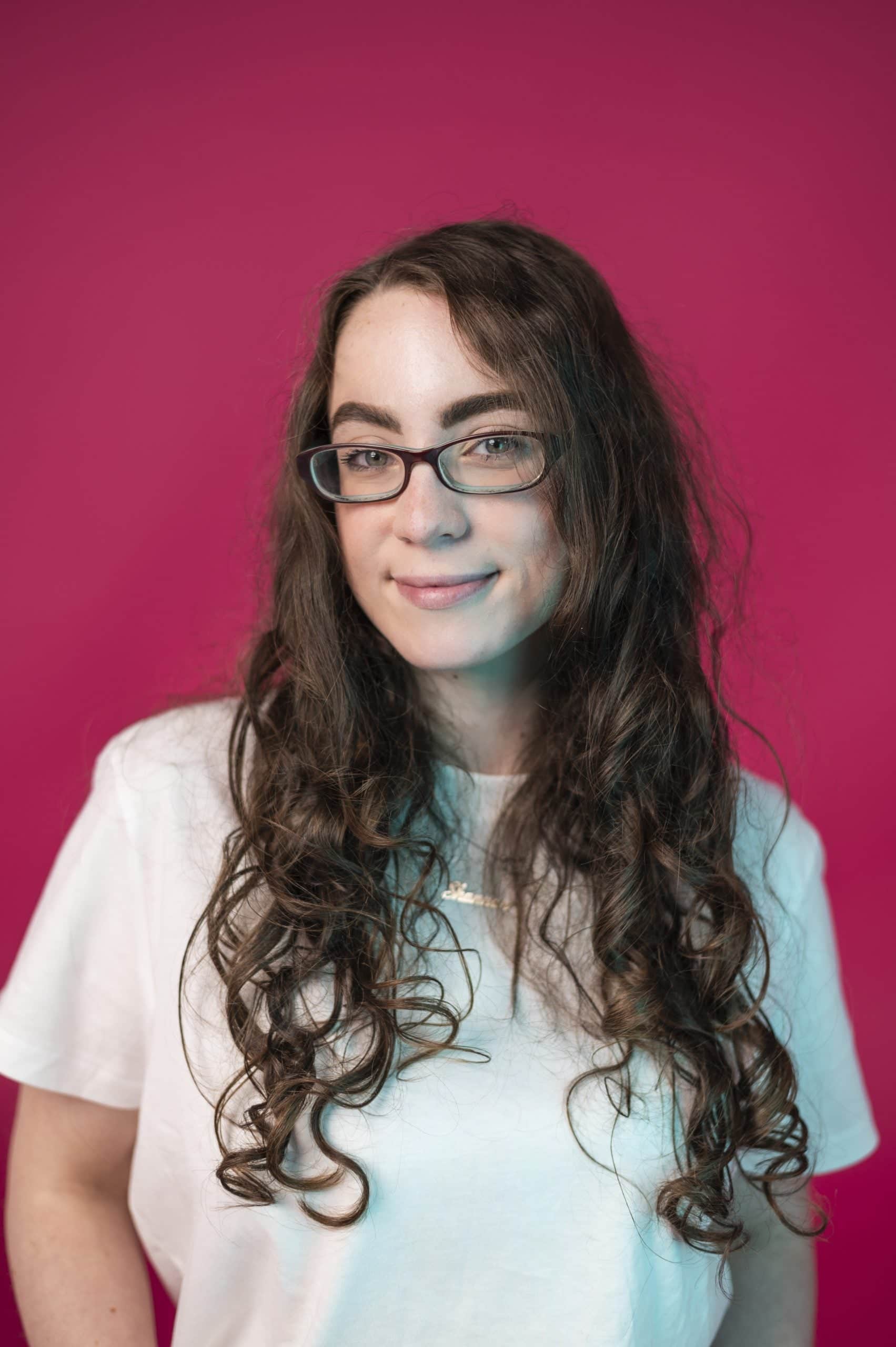 Woman with long curly hair and glasses against a pink background, wearing a white t-shirt.