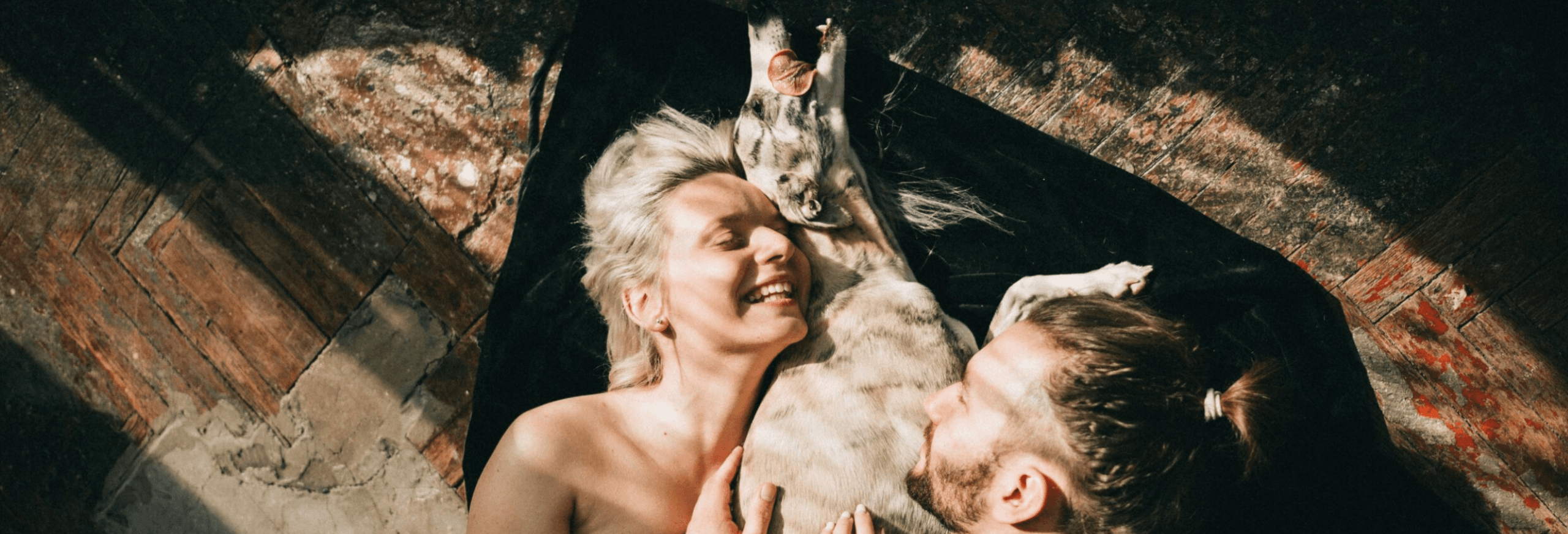 Couple lying down together gazing up at playful white dog overhead