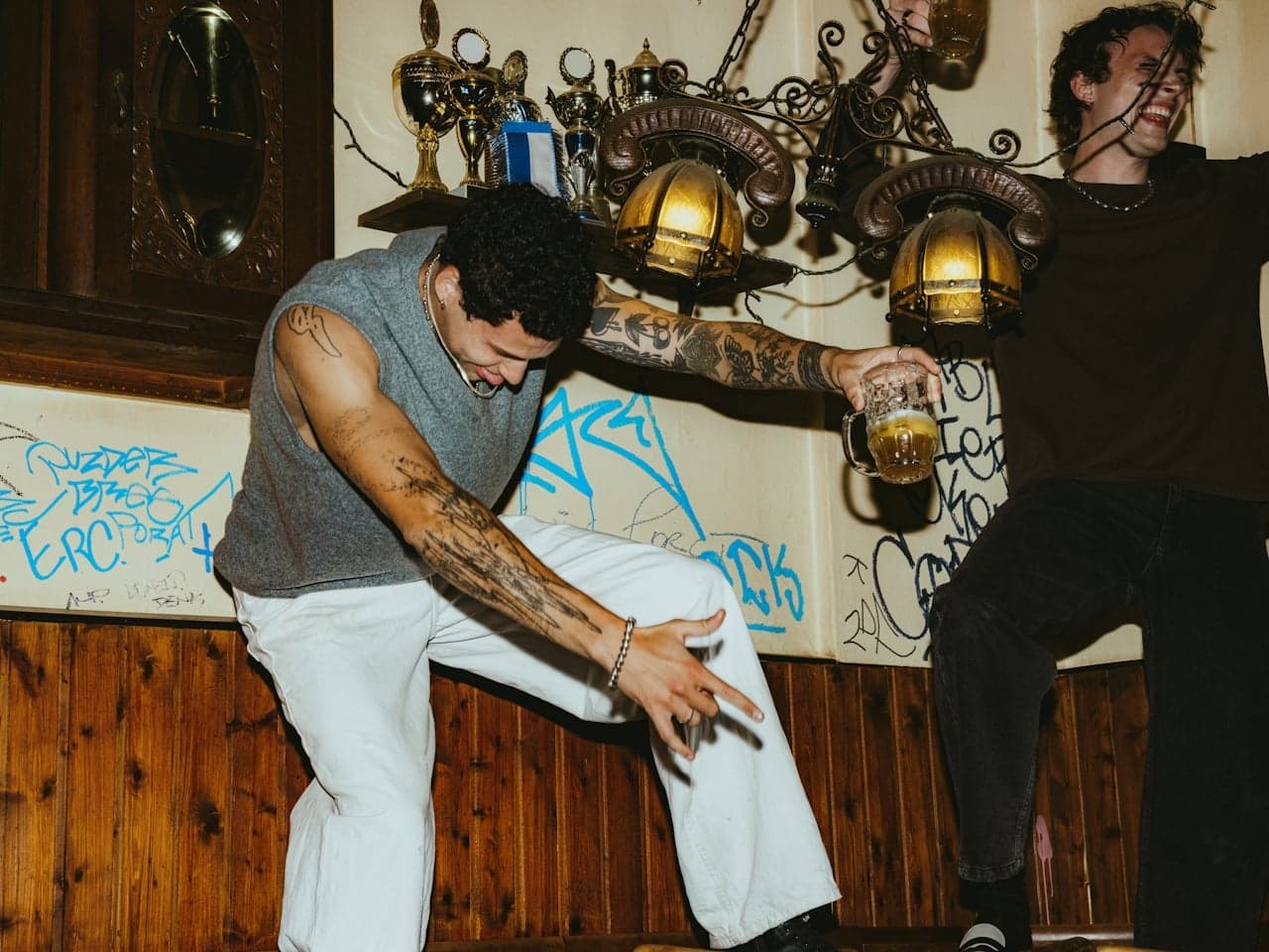Two men dancing on a wooden table in a lively bar with beer, graffiti, and trophies in the background.
