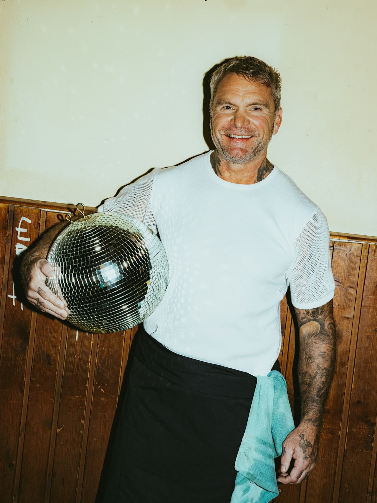 Man smiling, holding a disco ball, wearing a mesh white t-shirt and apron.
