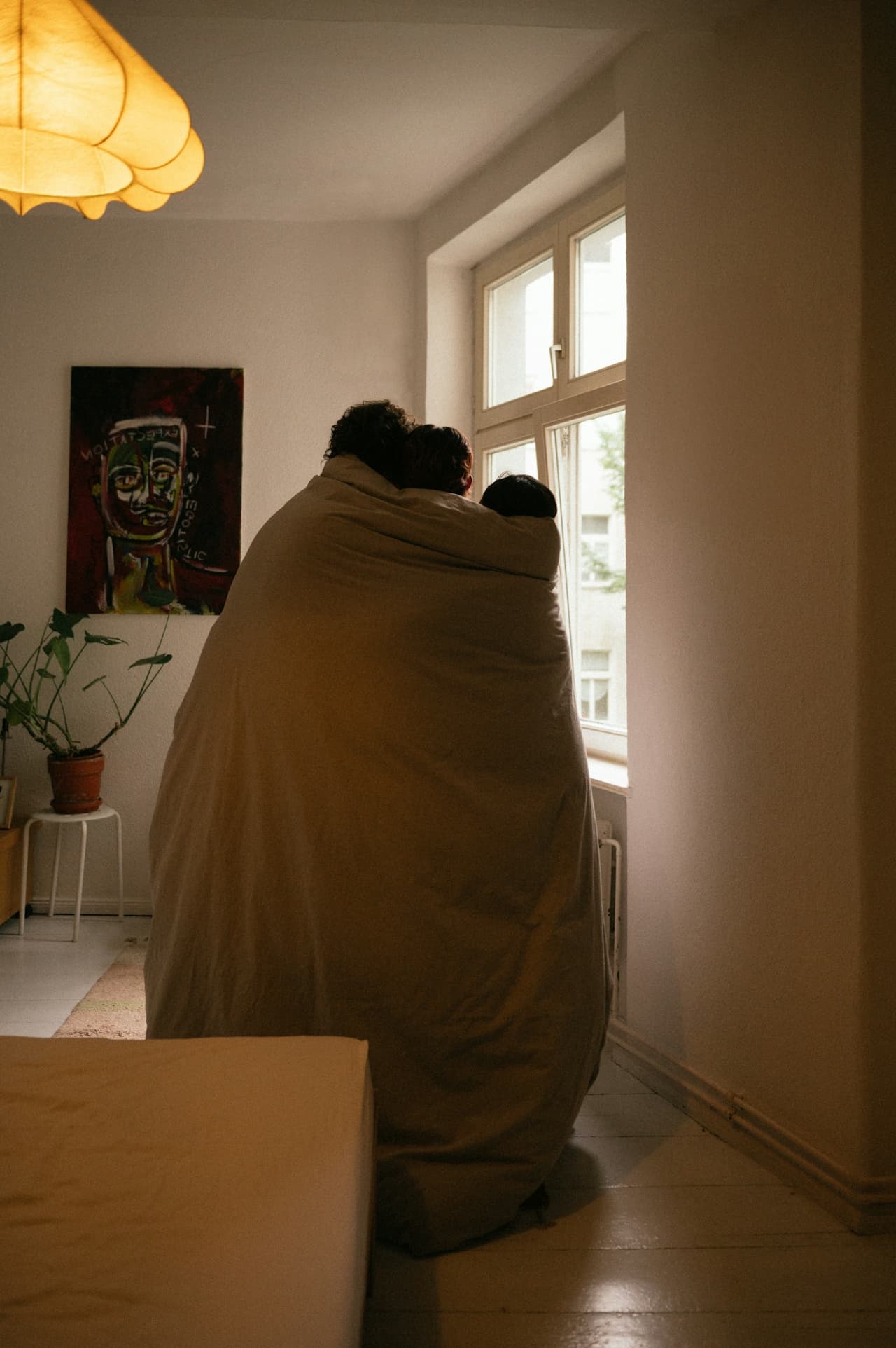 Two people wrapped in a blanket gaze out a window in a cozy, art-adorned living room.