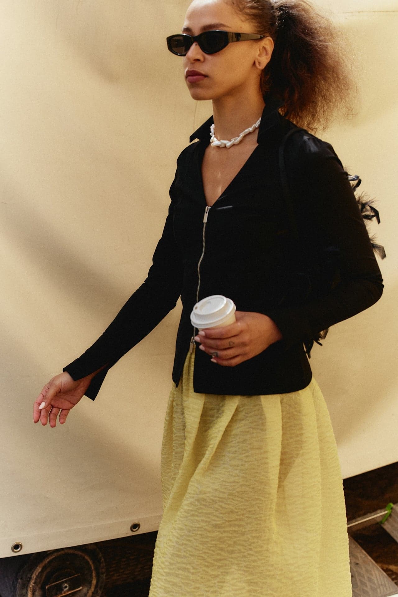 Woman in stylish black jacket and yellow skirt holding coffee cup, wearing sunglasses, reflecting urban fashion.