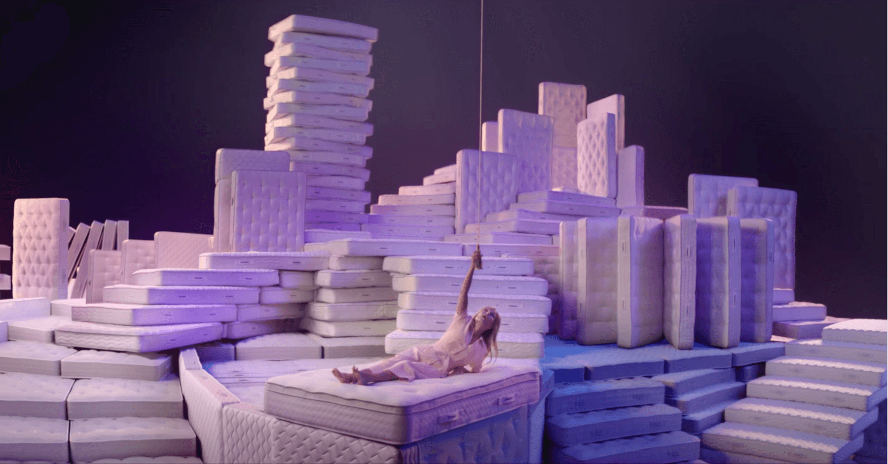 Person lounging on a mattress surrounded by stacked mattresses, showcasing mattress variety.