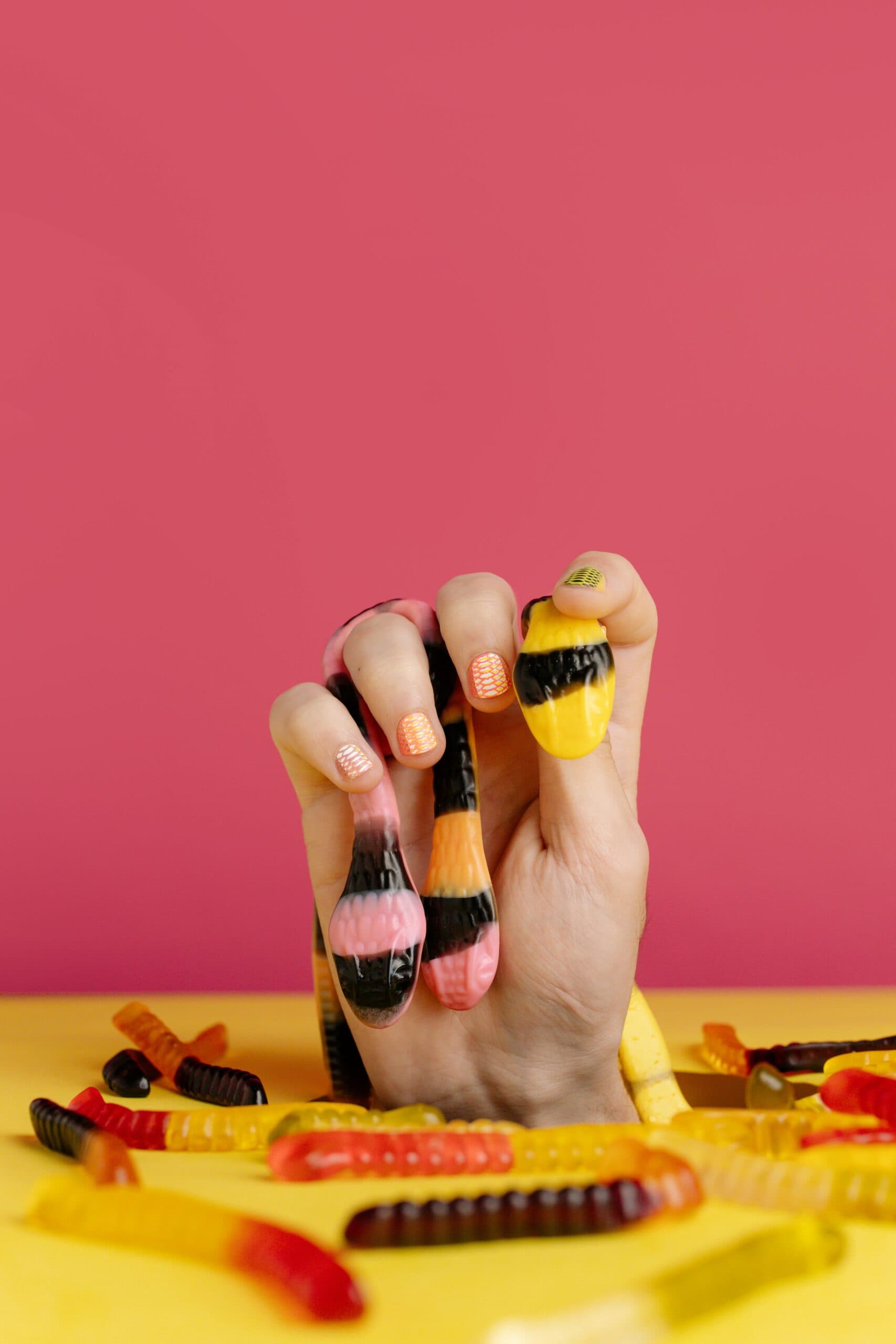 Hand holding gummy worm candies in creative candy arrangement with vibrant pink and yellow background