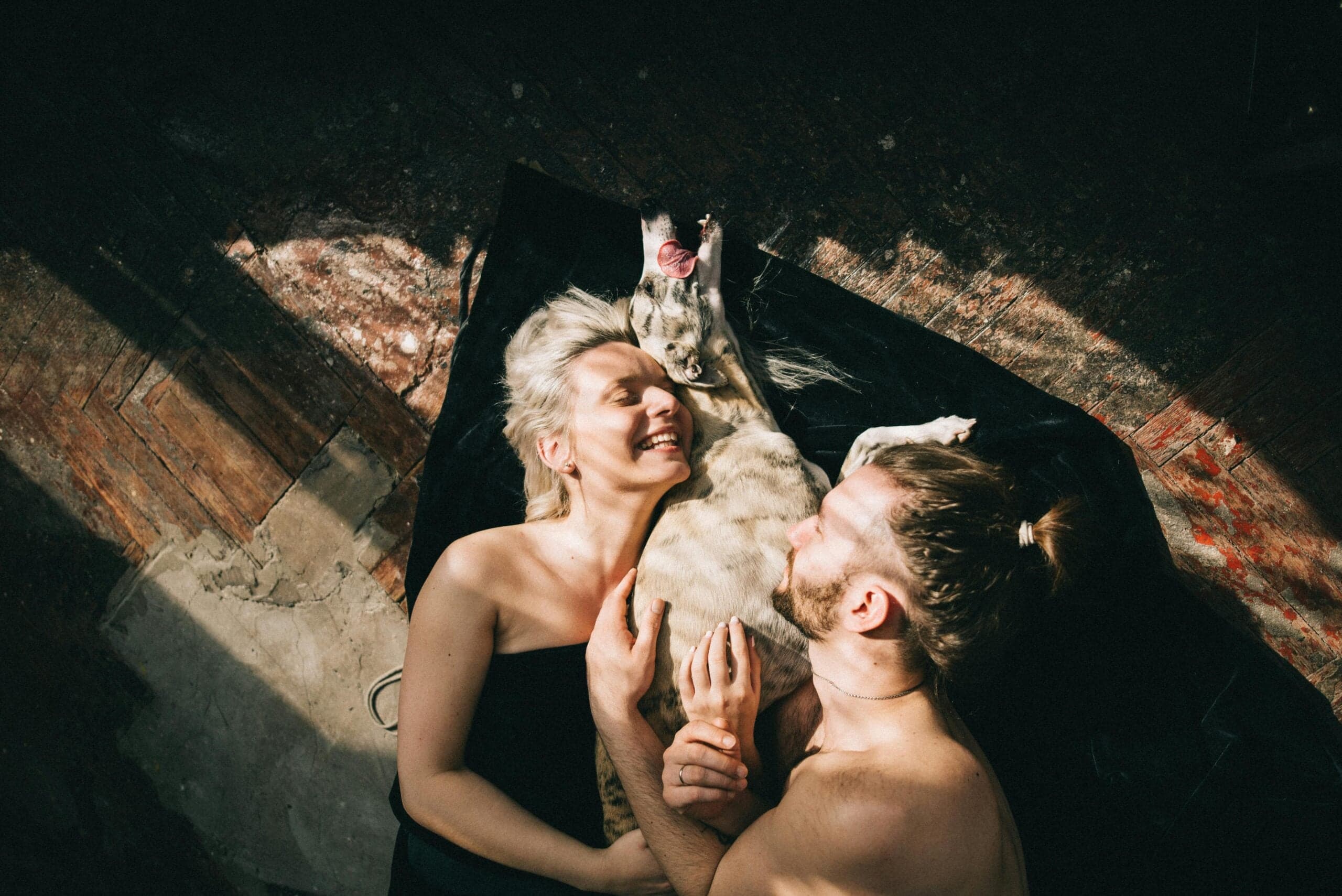 Couple lying on wooden floor holding white dog overhead, sharing joyful moment