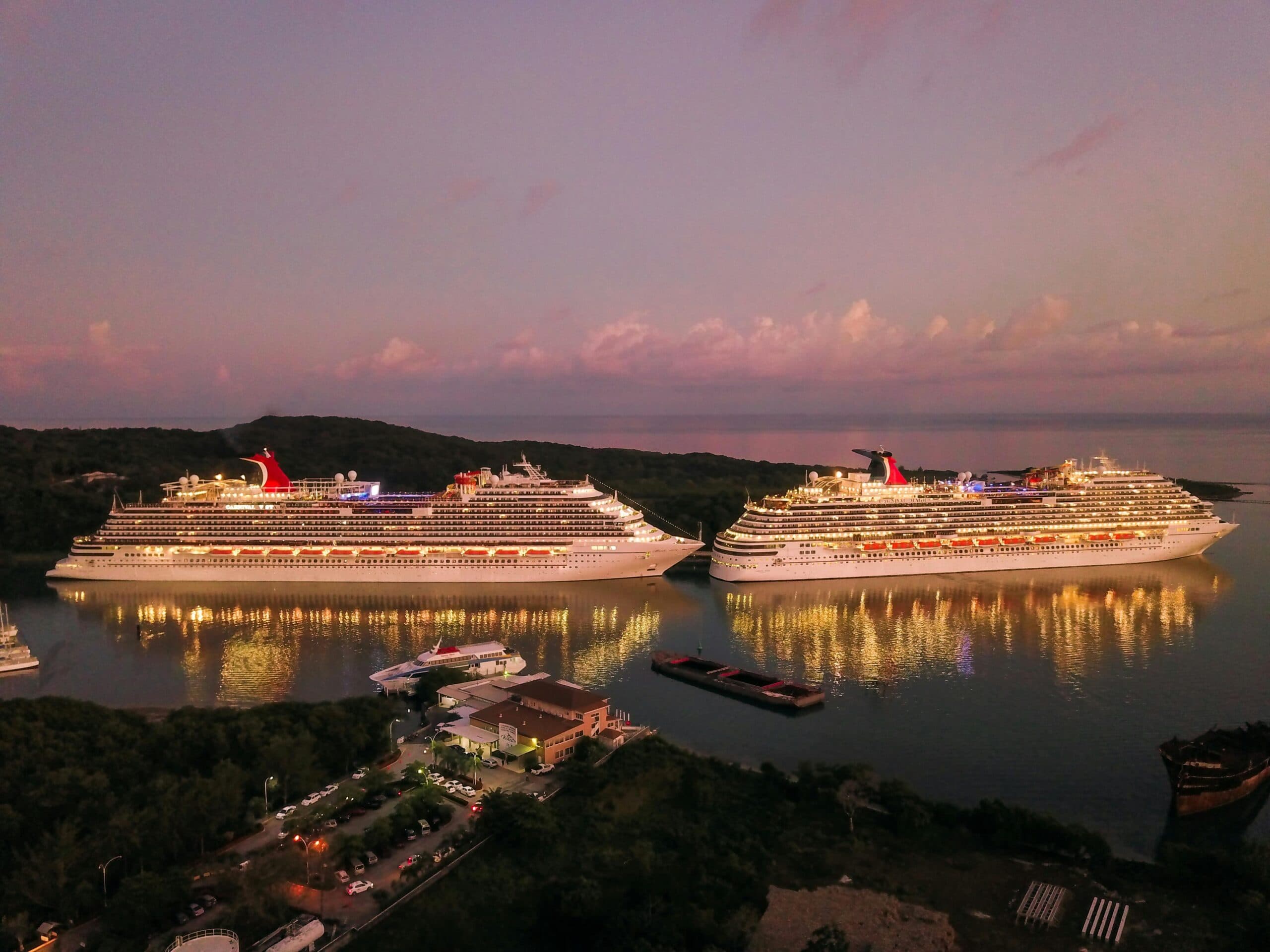 Two illuminated cruise ships docked at sunset near a forested coastline, reflecting on calm water.