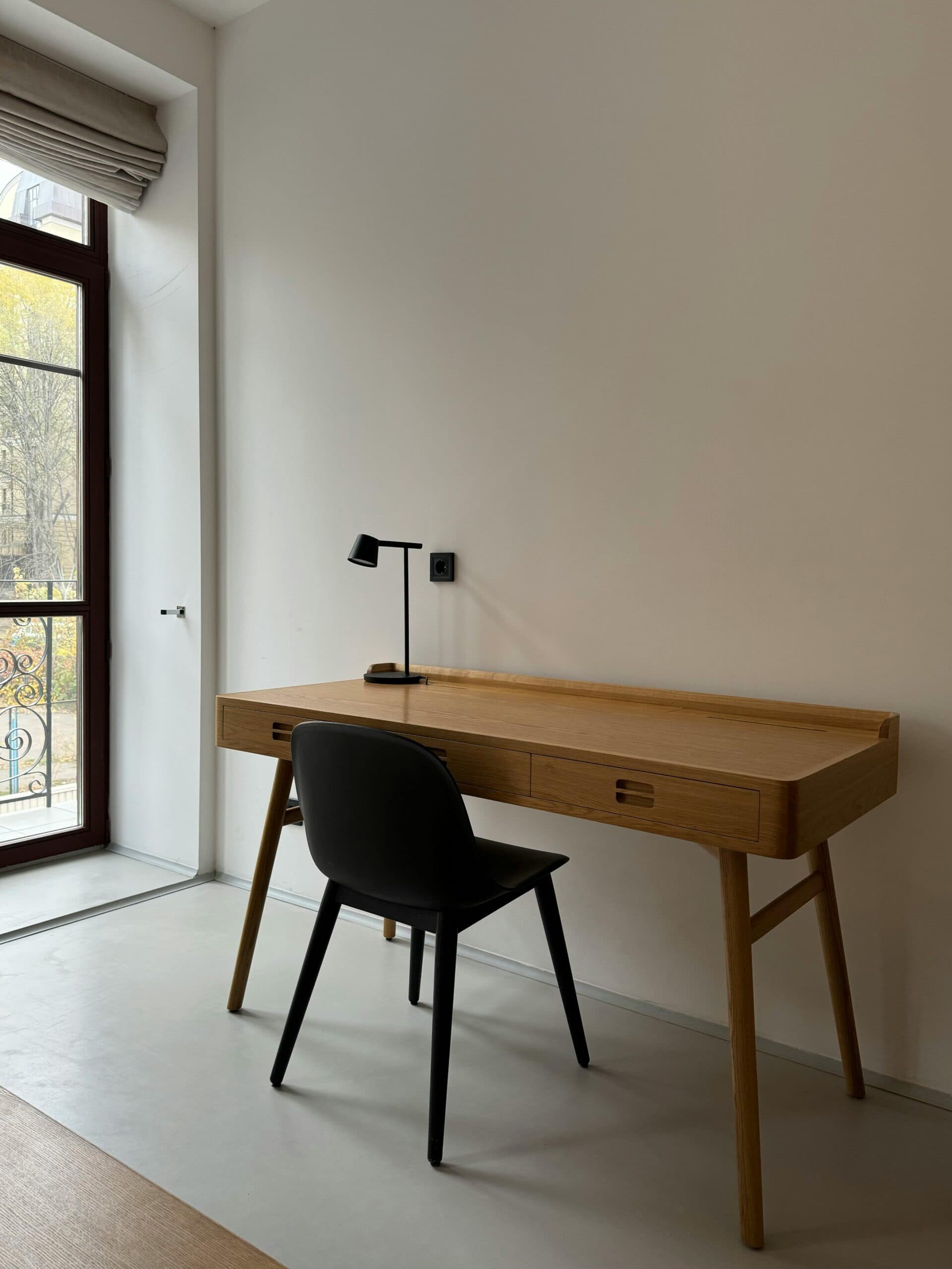 Minimalist home office workspace with wooden desk, black chair, and desk lamp by large window