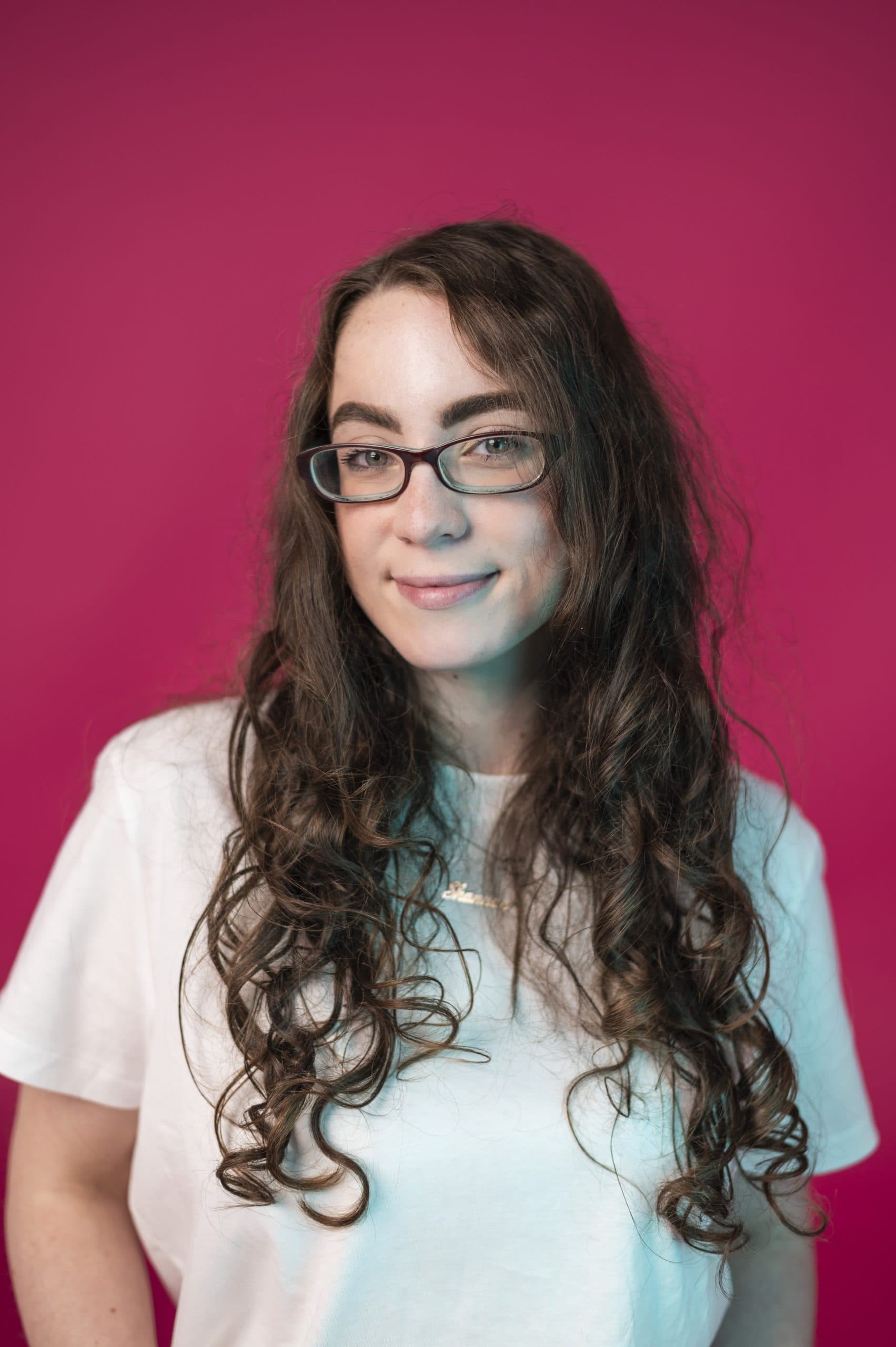 Woman with long curly hair and glasses against a pink background, wearing a white t-shirt.
