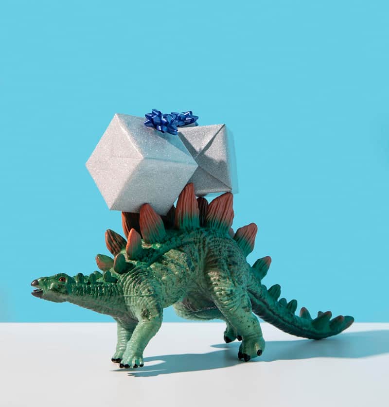 Toy stegosaurus carrying a silver gift box with a blue ribbon against a bright blue background.