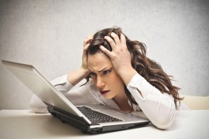 Frustrated women at laptop - her fingers are clasped against the side of her head and her brow is furrowed